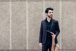 Manu Brazo with saxophone standing in front of concrete wall