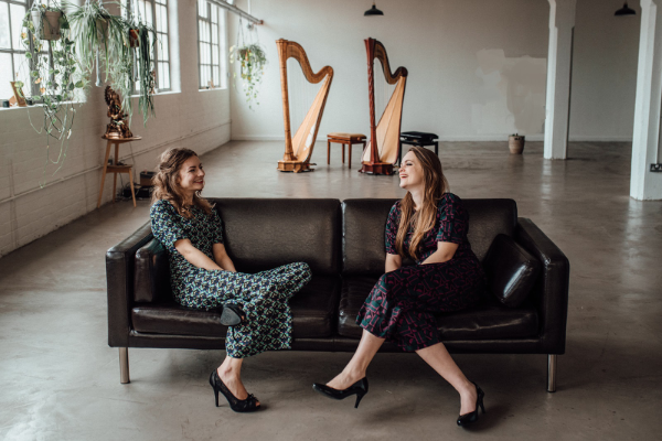 The Chroma Harp Duo sitting on a sofa with their harps in the backgound