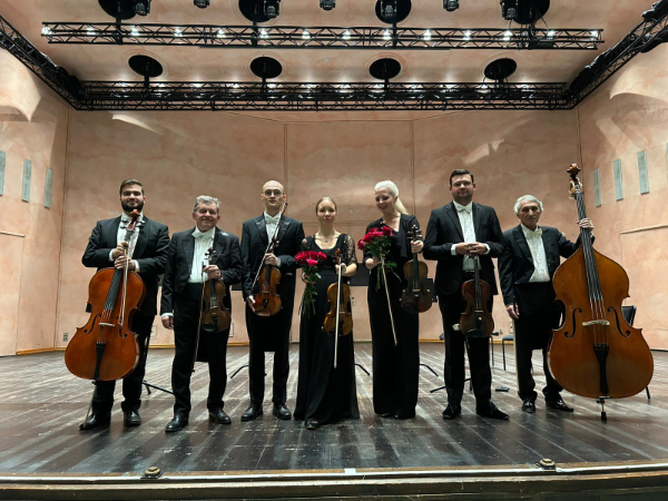 Seven performers from Chamber Philharmonic Europe standing, with their instruments, near the front of a stage
